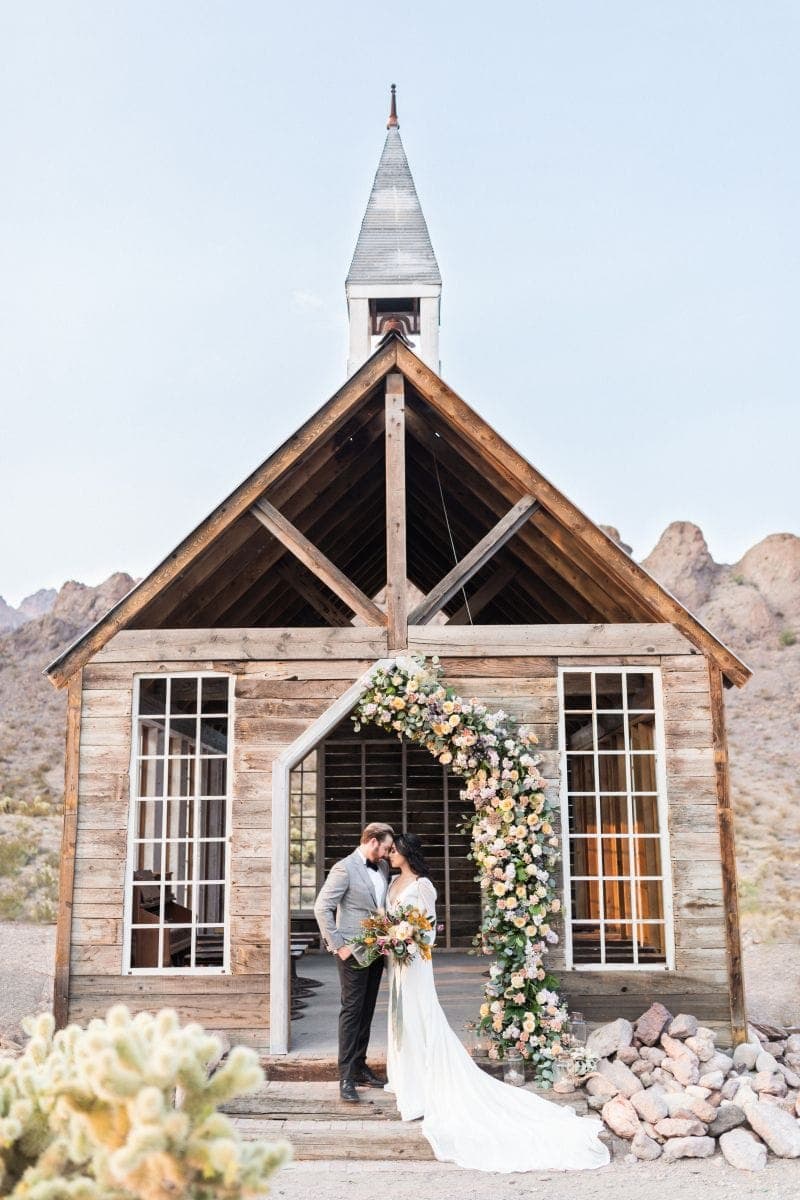 A couple embracing beneath a floral-draped chapel archway at Eldorado Canyon, a Scheme Events wedding