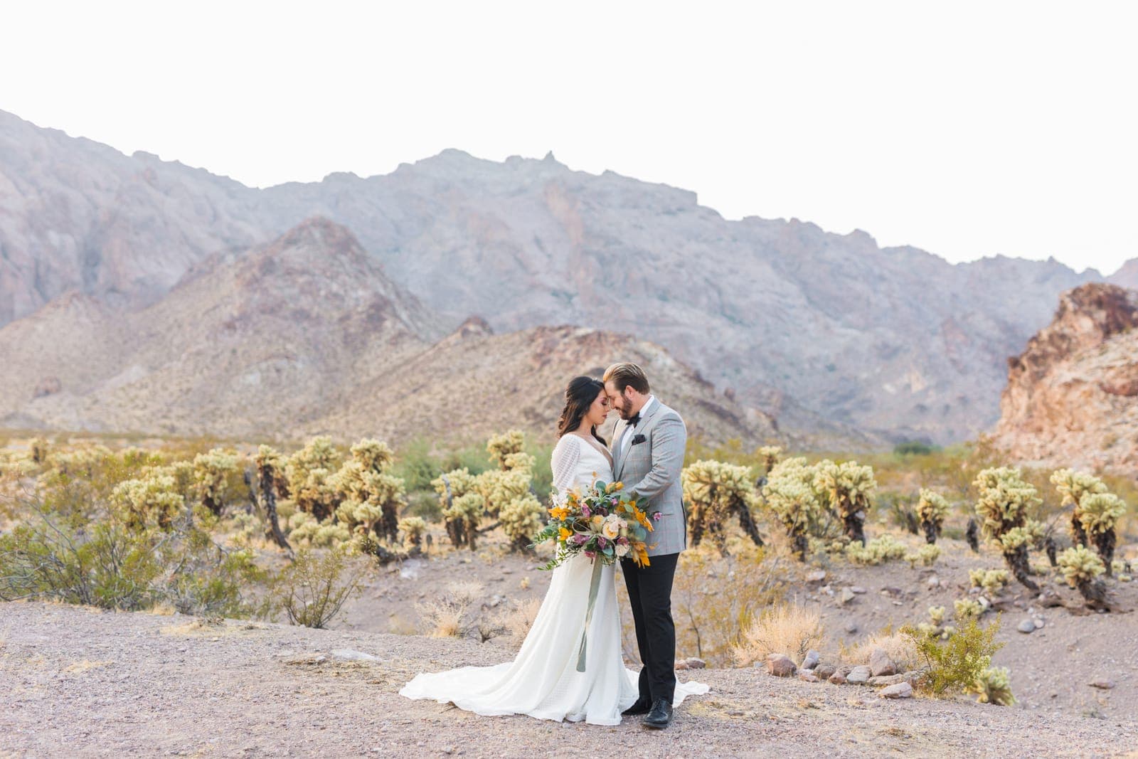 A couple embraces among cholla cactus with the Eldorado Canyon mountains behind them, from a Scheme Events elopement