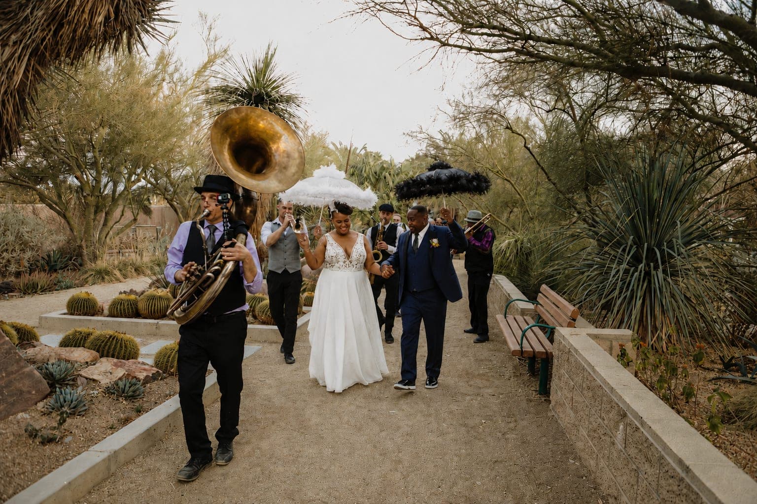 A brass band leads a couple under feather umbrellas in a New Orleans style second line through a desert garden, photography by New Romantix LV